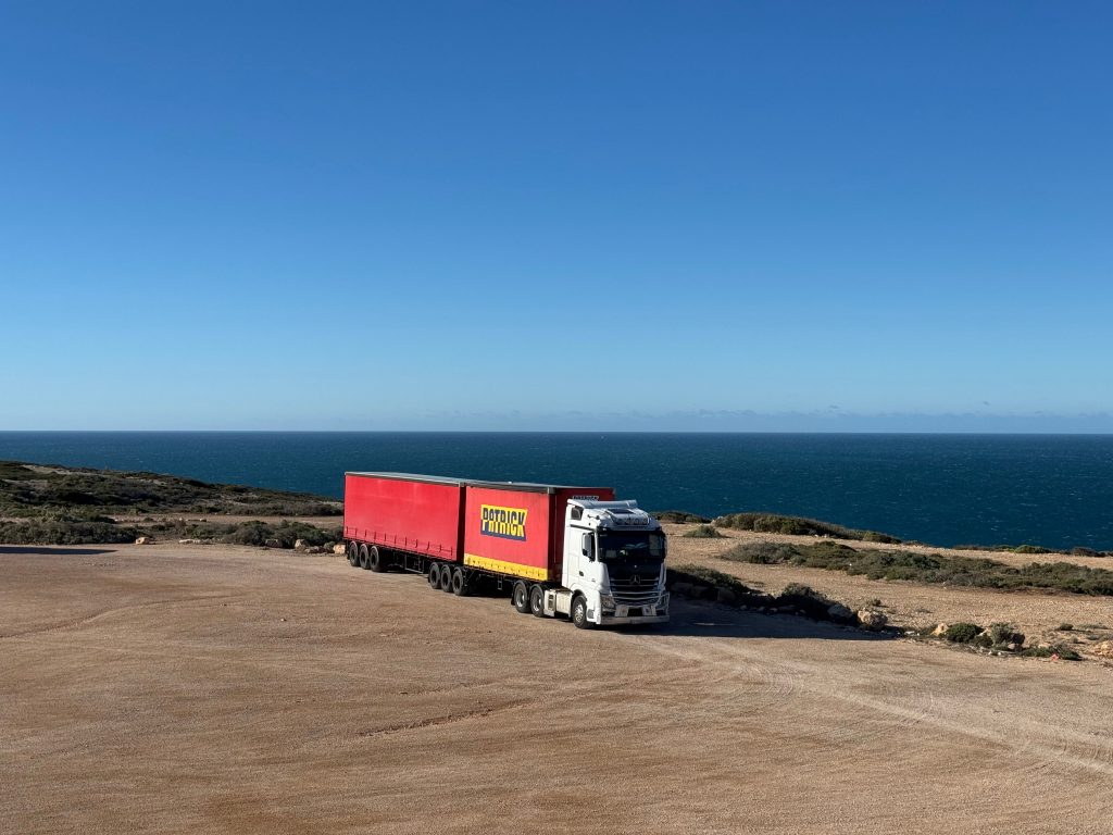 pexels-photo-34646235-34646235 A white truck with a red trailer travels on a coastal road next to the ocean.